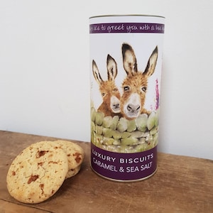 May include: A biscuit tin featuring a watercolour illustration of two donkeys. The tin is labelled "Luxury Biscuits Caramel & Sea Salt". Three caramel biscuits are in front of the tin on a wooden surface.
