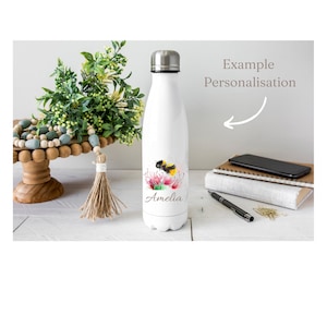 Puede incluir: Botella de agua de acero inoxidable blanca con un diseño de abeja y flor y el nombre Amelia impreso en ella. Ejemplo de personalización.