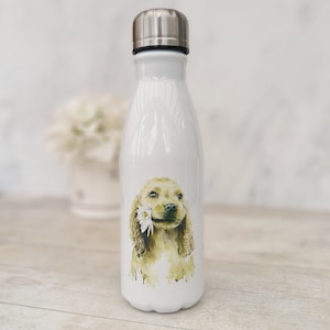 Puede incluir: Botella de agua de acero inoxidable blanca con una ilustración de acuarela de un cocker spaniel dorado sosteniendo una flor blanca en su boca.