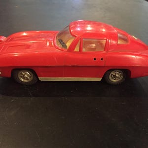 May include: A vintage red toy car with a sleek, aerodynamic design. The toy car has a clear windshield and side windows, gold-colored wheels, and a detailed front end. A classic collectible toy.