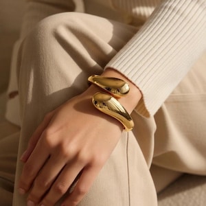 May include: A pair of gold-colored, curved bracelets. The bracelets are worn on a person's wrist, and the smooth, reflective surface of the bracelets catches the light. The bracelets have a modern, minimalist design.