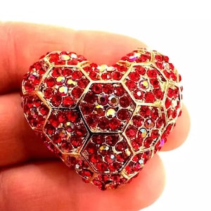 May include: A heart-shaped brooch featuring a gold-toned metal frame with a honeycomb pattern. Each hexagonal cell is filled with small, vibrant red crystals that catch the light, creating a textured and reflective surface across the entire piece.