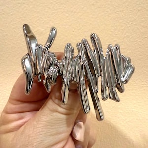 May include: A shiny, silver-colored ring with an abstract, flowing design. The ring features a series of irregular, elongated shapes that create a unique and modern aesthetic. The ring is held up against a neutral background.