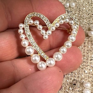 May include: A heart-shaped brooch featuring a gold-toned metal frame encrusted with clear rhinestones and accented with a row of white faux pearls. The design consists of an open heart structure with a pin mechanism on the back side.