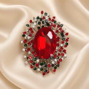 May include: A silver-toned brooch featuring a large, central oval-shaped red gemstone surrounded by a decorative, branch-like metal frame encrusted with small, round red, green, and clear crystals arranged in a dense, festive, floral-inspired pattern.