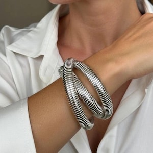 May include: A silver-colored, ribbed bracelet is wrapped around a wrist. The bracelet has a flexible, coiled design. The person is wearing a white button-down shirt. The bracelet is a statement piece.