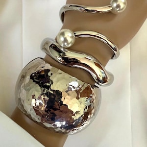 May include: A collection of silver-toned bracelets. The bracelets include a hammered metal cuff, a smooth, curved bangle, and two open-ended bangles, each with a large, round pearl accent. The jewelry is displayed on a wrist.