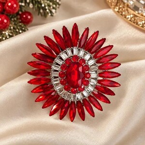 May include: A circular brooch featuring a large central oval red gemstone surrounded by a ring of smaller round red stones, a border of clear baguette-cut crystals, and an outer layer of elongated marquise-cut red crystals in a sunburst pattern.