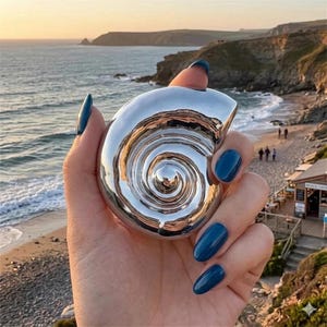 May include: A polished silver seashell-shaped object with a spiral design. The object is held in a hand with blue nail polish, reflecting the beach and ocean in its surface. The background shows a beach scene.