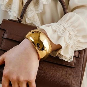 May include: A wide, polished gold-tone cuff bracelet with a smooth, reflective surface. The bracelet is worn on the wrist, positioned against a dark brown textured handbag and a cream-colored long-sleeved garment with delicate lace trim at the cuffs.