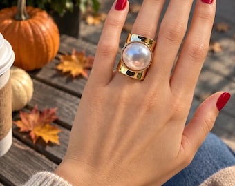 May include: A wide gold-tone ring featuring a large, round, iridescent white pearl set in the center. The band has a smooth, polished metallic finish that wraps around the finger, creating a bold and prominent statement piece of jewelry.