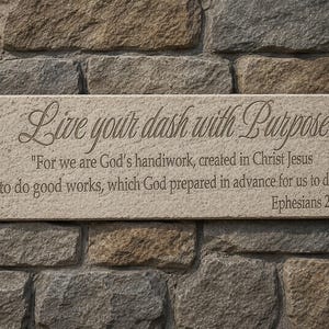 May include: A rectangular stone plaque with the inscription "Live your dash with Purpose." Below, it reads, "For we are God's handiwork...Ephesians 2:10." The plaque is light beige and set against a textured stone wall.