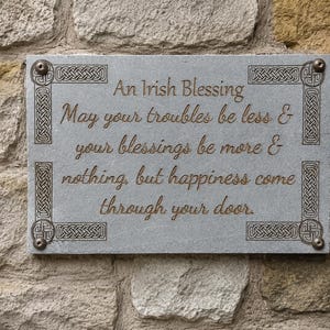 May include: A rectangular gray stone plaque with an Irish blessing engraved in brown script. The plaque is adorned with Celtic knotwork in the corners and is mounted on a stone wall. The text reads: "An Irish Blessing..."
