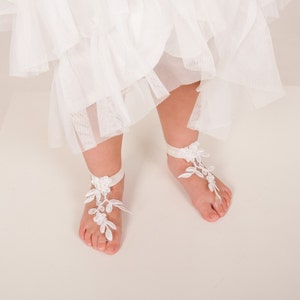 Puede incluir: Sandalias de encaje blanco para pies descalzos con un delicado dise&ntilde;o floral. Las sandalias se atan con una cinta blanca alrededor del tobillo.