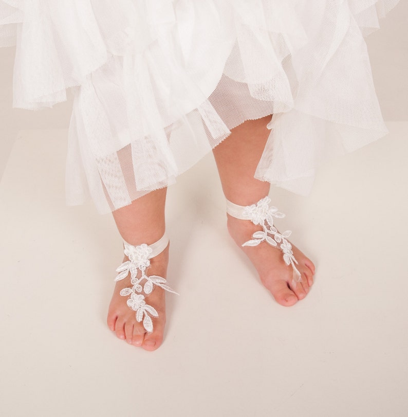 Puede incluir: Sandalias de encaje blanco para pies descalzos con un delicado dise&ntilde;o floral. Las sandalias se atan con una cinta blanca alrededor del tobillo.