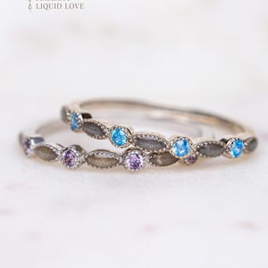 May include: Two silver rings with alternating blue and purple gemstones. The rings have a decorative design with oval-shaped accents. The rings are set against a soft, light background. The text "MAMMA'S LIQUID LOVE" is in the upper left corner.