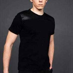 Puede incluir: Una camiseta negra de manga corta con un panel de cuero negro en el hombro.