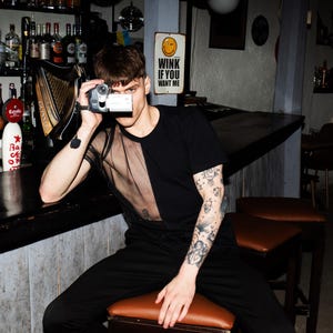 Puede incluir: Una persona lleva una camiseta negra con un panel de malla transparente y una camiseta negra debajo. Está sosteniendo una cámara de video vintage y mirando a la cámara. La persona tiene tatuajes en el brazo y está sentada en un bar con un letrero que dice "Wink if you want me".
