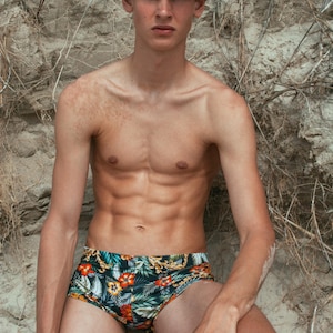 Puede incluir: Ba&ntilde;ador con estampado floral tropical en tonos verdes, naranjas y dorados. El ba&ntilde;ador presenta un fondo negro y un corte cl&aacute;sico. El modelo est&aacute; sin camisa, con tez bronceada, y est&aacute; sentado contra un fondo arenoso.