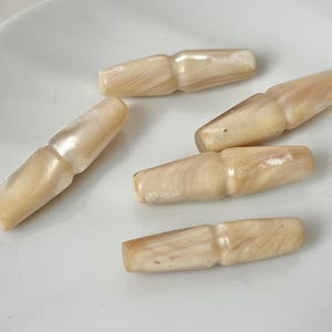 Peut inclure: Six fermoirs à bascule beige clair avec un éclat nacré sont disposés sur une surface blanche. Les fermoirs ont une forme cylindrique avec un centre légèrement plus large et des extrémités arrondies.
