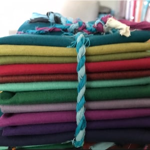 May include: A stack of fabric scraps in various colors, including teal, green, red, purple, and brown. The fabric is tied together with a blue and white braided rope.