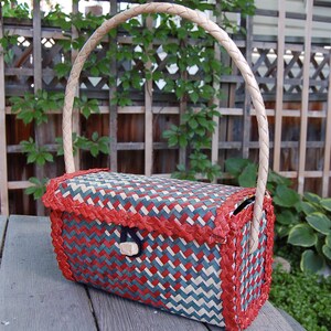 Op de afbeelding: Een geweven mandtas met een rood, wit en blauw geometrisch patroon. De handgreep is gemaakt van lichtbruin materiaal.