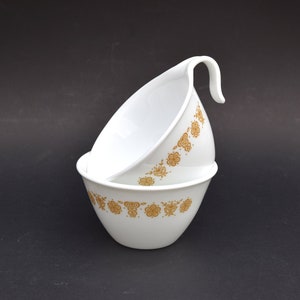 Puede incluir: Dos cuencos de cerámica blanca con un patrón floral dorado. El cuenco de arriba tiene un asa.