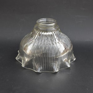 May include: A clear glass lamp shade featuring a bell-shaped silhouette with vertical ribbed detailing and a ruffled, scalloped edge. The top opening has a threaded fitter neck designed for secure attachment to a light fixture base.