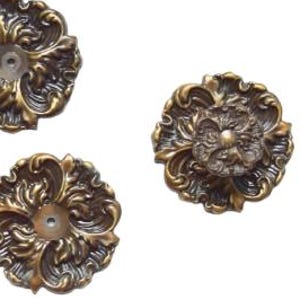 May include: Four round metal drawer knobs with an antique bronze finish and intricate, raised floral scrollwork patterns. Three of the knobs feature a central circular hole, while one knob is attached to a small metal mounting screw.