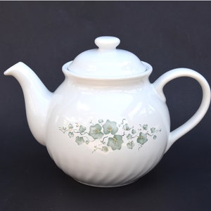 Op de afbeelding: Witte theepot van keramiek met een groen en geel bloemenmotief. De theepot heeft een deksel en een handvat.