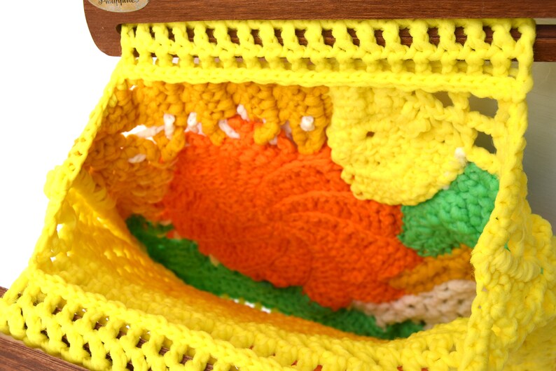 Pu&ograve; includere: Un pezzo all'uncinetto fatto a mano, colorato, con un vivace mix di filati gialli, arancioni, verdi e bianchi. Il pezzo &egrave; sospeso a un telaio di legno, mostrando un design strutturato e a trama aperta. L'estetica generale ricorda l'artigianato vintage.
