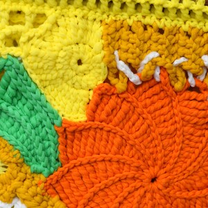 Pu&ograve; includere: Primo piano di un colorato pezzo all'uncinetto fatto a mano. Il design presenta grandi motivi floreali strutturati nelle tonalit&agrave; del giallo, dell'arancione e del verde. Le cuciture bianche accentuano i bordi, creando un tessuto vibrante e tattile.