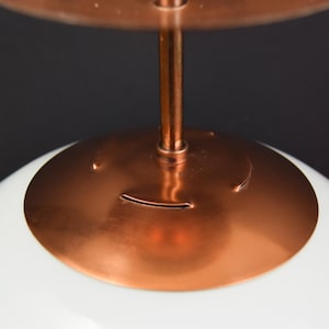 May include: Close-up of a modern pendant light. It has a white, rounded glass shade, a copper-coloured metal canopy and stem. The design is minimalist and elegant, suitable for various interior styles.