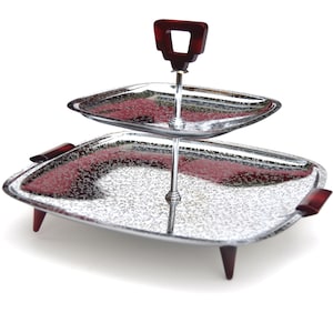May include: Two-tiered silver serving tray with a red plastic handle. The tray has a textured surface and stands on four legs.