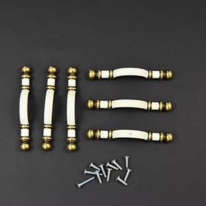 May include: Set of six white and brass cabinet knobs. The knobs are shaped like a rounded rectangle with a slight curve. The knobs are attached to screws.
