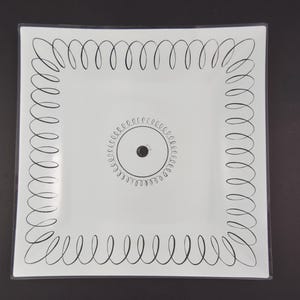 Può includere: Piatto quadrato bianco con un design a spirale nero sui bordi e un motivo circolare al centro. Il piatto ha una finitura lucida ed è posto su uno sfondo scuro.