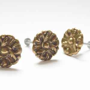 Puede incluir: Tres tiradores de armario de latón de estilo antiguo con diseños florales ornamentados. Cada tirador presenta un patrón en relieve detallado y está montado sobre un tornillo plateado. Los tiradores están sobre un fondo blanco.