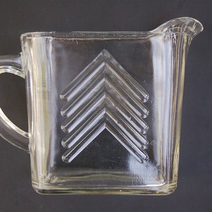May include: A clear glass jug with a geometric design of overlapping arrows. The jug has a handle and a wide mouth.