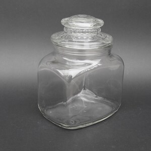 Op de afbeelding: Een glazen pot met een vierkante body en een glazen deksel. Het deksel heeft een verhoogde rand en een gladde, afgeronde bovenkant.