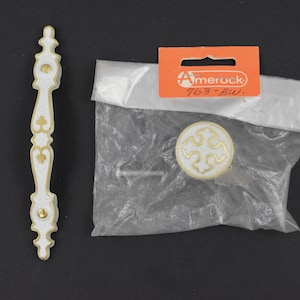 May include: White cabinet knob with a gold trim and a decorative cross design. The knob is packaged in a plastic bag with a label that reads "Amerock 763-BW."
