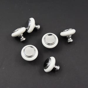 May include: Six white ceramic cabinet knobs with a silver metal trim and a silver screw. The knobs are round in shape with a slightly raised centre.