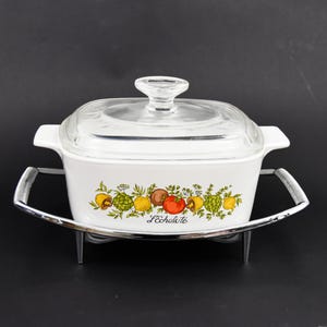 Puede incluir: Plato para hornear blanco con tapa de vidrio y un diseño floral con hojas verdes, limones amarillos, tomates rojos y la palabra "Lechalite". El plato está sobre un soporte de metal plateado.