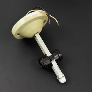 Puede incluir: Una antigua lámpara de techo con una base de color crema y un brazo plateado. La lámpara tiene cables negros y blancos que salen de la base y un componente cilíndrico negro. La lámpara está diseñada para cableado eléctrico.