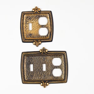 May include: Two decorative light switch covers. The covers have a gold and black finish with an ornate, textured design. Each cover has a decorative fleur-de-lis at the top and bottom. One cover has two outlets and a switch, the other has one switch and two outlets.