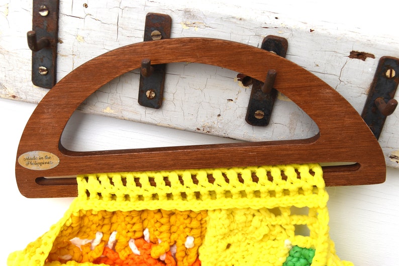 Pu&ograve; includere: Un telaio per borsa in legno con una parte superiore semicircolare e una base rettangolare, che contiene una borsa all'uncinetto colorata. La borsa presenta sezioni gialle, arancioni e verdi. Il telaio ha una piccola etichetta che dice "made in the Philippines".