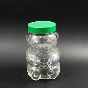 Può includere: Un barattolo di vetro trasparente a forma di orso con un coperchio di plastica verde. Il barattolo è vuoto e ha una superficie testurizzata.