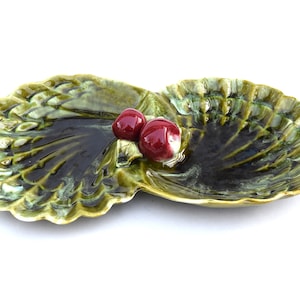 Pode incluir: Um prato de servir de cerâmica verde em forma de duas conchas com bagas de cerâmica vermelha no topo.