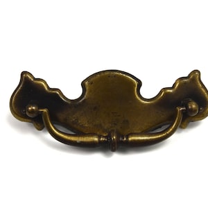 May include: A brass drawer pull with a decorative, ornate design. The pull has a curved handle and a central, flat plate with a raised edge.