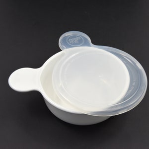 May include: A white baby bowl with a clear lid. The bowl has a handle on each side. The lid is transparent and has text printed on it. The bowl is on a black surface.