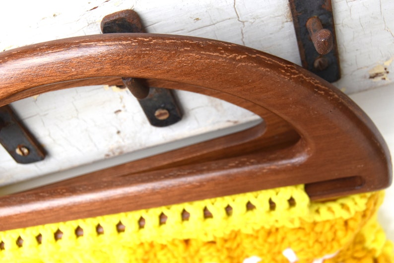 Pu&ograve; includere: Primo piano di un manico di borsa vintage in legno. Il manico &egrave; di colore marrone scuro e ha un design curvo e arcuato. Il tessuto all'uncinetto giallo e bianco della borsa &egrave; visibile sotto.